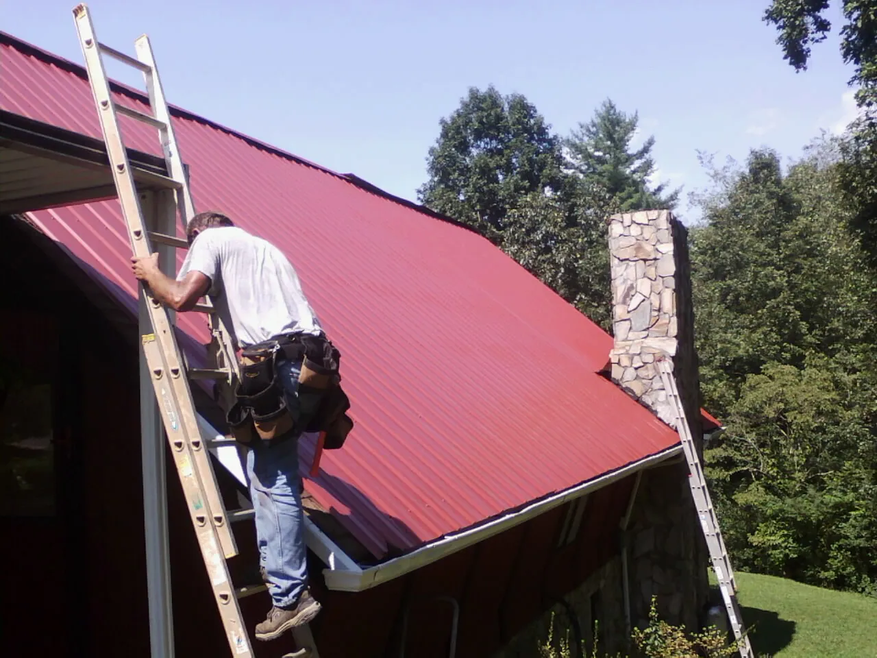Roofer on a ladder beside a red metal roof and stone chimney — Soffit & Fascia Repair by Roofing Brothers in Worton